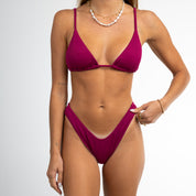 A woman models the Honey Swim Berry Ribbed Triangle Set bikini with adjustable straps, accessorized with layered necklaces and a bracelet. Her face is not shown as she poses against a white background.
