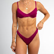 A woman wears the Honey Swim Berry Ribbed Underwire Top bikini with gold jewelry, posing against a white background, highlighting its supportive fit and ultra-soft ribbed fabric.