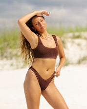 A woman in the Honey Swim Brown Luxe Set stands on a sandy beach with green grass and a cloudy sky, shielding her eyes from the sun with one arm.