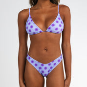 A woman models the Honey Swim Daybreak Triangle Set, a light purple bikini with starburst prints and adjustable straps, standing against a plain white background with her arms relaxed by her sides.