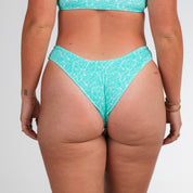A person is seen from the back wearing Honey Swim’s Drift Ribbed | Cheeky Bottoms in a light blue pattern. They have tattoos on their left arm and wrist, and gold bracelets on the right, against a plain white background.