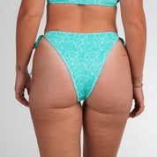 A person shown from the back wears Honey Swims Drift Ribbed String Bottoms in a tonal green-on-green print, featuring side ties, gold bracelets, and a small wrist tattoo.