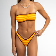 A woman models the Honey Swim Gold Coast Ribbed Cinched Bandeau Set bikini against a white background, holding the side of her bikini bottom. She has long hair and accessorizes with necklaces and bracelets.