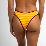Someone wearing Honey Swim’s Gold Coast Ribbed Bottoms in yellow and orange stripes with dark trim, viewed from behind against a plain light background, showing only their lower back and upper legs.