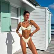 A woman in a Honey Swim Cream Ribbed Underwire Set and a shell necklace stands smiling in front of a white beach house with mint-green shutters, embodying perfect summer days. The ocean and blue sky serve as the idyllic backdrop.