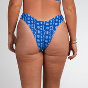 A person faces away from the camera in Honey Swim’s Lagoon Ribbed | Cheeky Bottoms, featuring vibrant blue with white abstract patterns. Their sun-kissed look is complemented by bracelets and small tattoos.