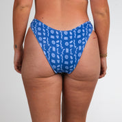 A person stands facing away, wearing Honey Swims Lagoon Ribbed | Classic Bottoms. The ribbed texture adds interest, while small tattoos and a gold bracelet accent the relaxed pose against a plain white background.