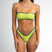 A woman models the Honey Swim Leo Cinched Bandeau Top, a green geometric print bikini with blue trim, against a white background. She has long brown hair and minimal jewelry, hands relaxed at her sides.