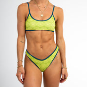 A woman models the Honey Swim Leo Bralette top, a yellow patterned bikini with blue trim, against a white background. She accessorizes with bracelets and necklaces; her face is not visible.