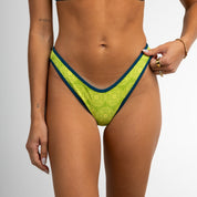 A woman models the Honey Swim Leo | Classic Bottoms, featuring a green and blue geometric print, posing against a white background with her left hand by her side and right hand lightly holding the waist.
