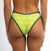 A person is shown from the back modeling Honey Swim’s Leo | Classic Bottoms, a green bikini with a geometric print and bold contrast detail, against a plain white background.