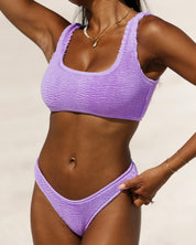A woman stands on a sandy beach wearing the Honey Swim Lilac Luxe Top, her gold jewelry and sun-kissed skin enhancing the luxe vibe against the blurred shoreline backdrop.