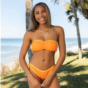 A woman stands by the beach, smiling in a Honey Swim Marigold Terry Cinched Bandeau Set, with palm trees and ocean waves behind her under the sun—ideal beach day attire.