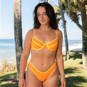 A woman with long brown hair stands by palm trees and the beach, wearing the Honey Swim Marigold Terry Classic Underwire Set, looking at the camera against an ocean and blue sky backdrop.