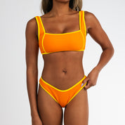 A woman models the Honey Swim Marigold Terry Squareneck Set—a textured, bright orange bikini with yellow trim. Cropped at shoulders and thighs, she holds the bikini bottom against a plain white background.