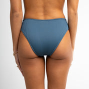 A person is seen from the back wearing Honey Swim’s Midnight Ribbed | Bottoms - All Styles, which are high-waisted and offer full coverage. Their arms rest at their sides, showing a bracelet on the left wrist against a plain white background.