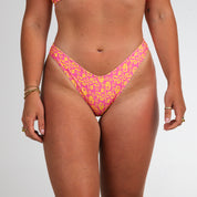 A person models Honey Swim’s Wildflower | Classic Bottoms in a pink and yellow pattern, featuring a pop of pink and visible tan lines, standing against a plain white background.