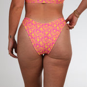 A person wears Honey Swims Wildflower | Classic Bottoms bikini, featuring pink and yellow hues. Their hand with gold bracelets rests on their hip, set against a plain white background.