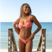 A woman in the Honey Swim Wildflower Ruched Underwire Set stands smiling on a wooden boardwalk at the beach, with the ocean and blue sky in the background.