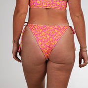 A person faces away from the camera, wearing Honey Swim’s Wildflower | String Bottoms in pink and yellow floral print with adjustable side ties, arms relaxed by their sides, against a plain white backdrop.