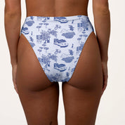 Someone is wearing Honey Swims Amalfi High Waisted Bottoms featuring a blue and white pattern of bicycles, houses, trees, and boats. These bottoms offer moderate coverage and a supportive fit against a plain white background.