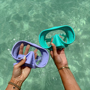 Two hands hold Honey Swims Honey Goggles, one lavender and one teal, above clear turquoise water. Sunlight dances on rippling water, highlighting painted nails and bracelets with a snug, leak-proof fit.