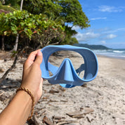 A hand holding Honey Goggles by Honey Swim frames a sandy beach with driftwood. Green trees line the beach, and the ocean rests under a clear blue sky.