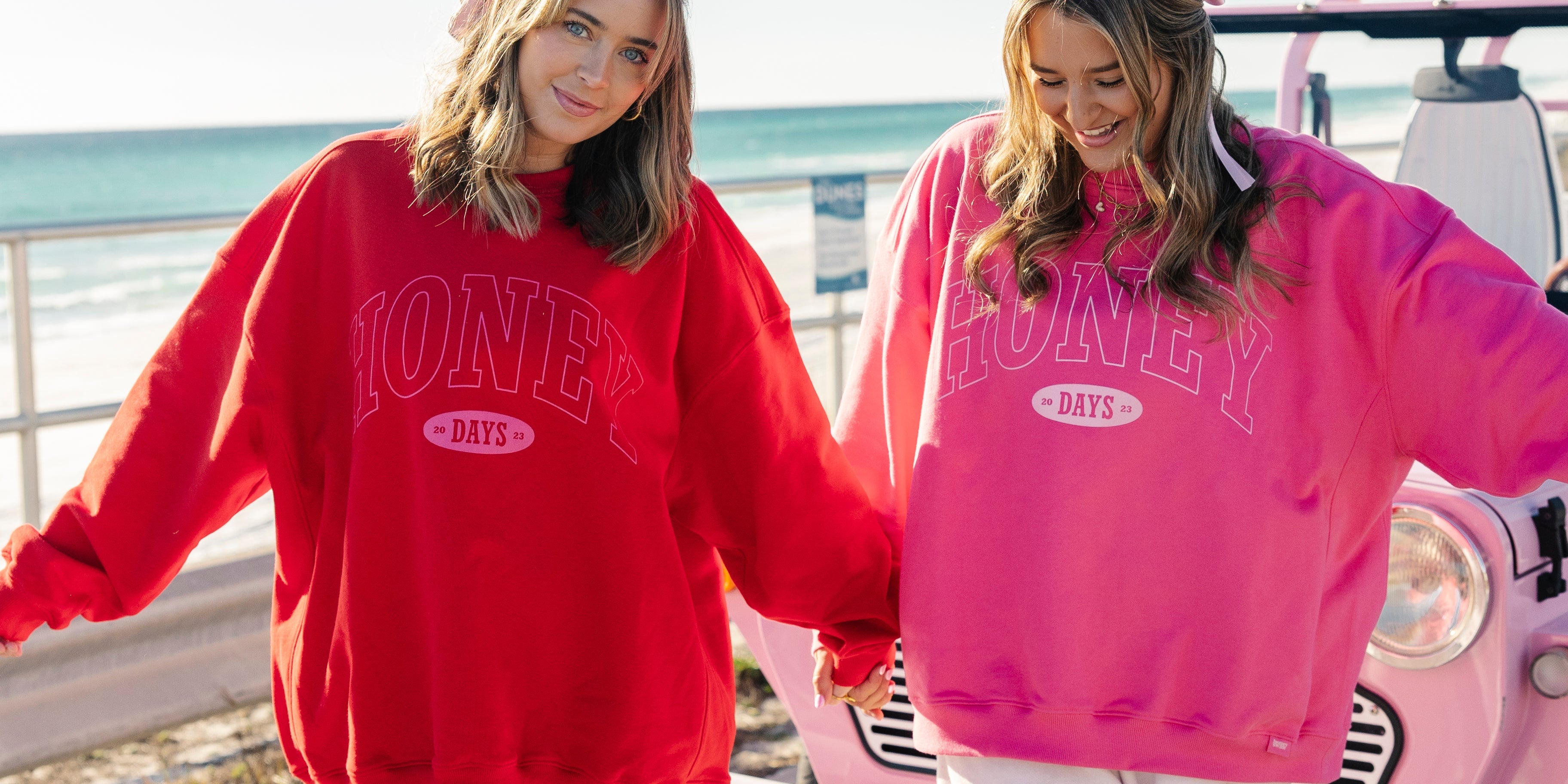 Two women smiling and holding hands by the ocean, wearing matching oversized sweatshirts—one red, one pink—with “HONEY DAYS” text, white pants, and standing beside a pink car on a sunny day.