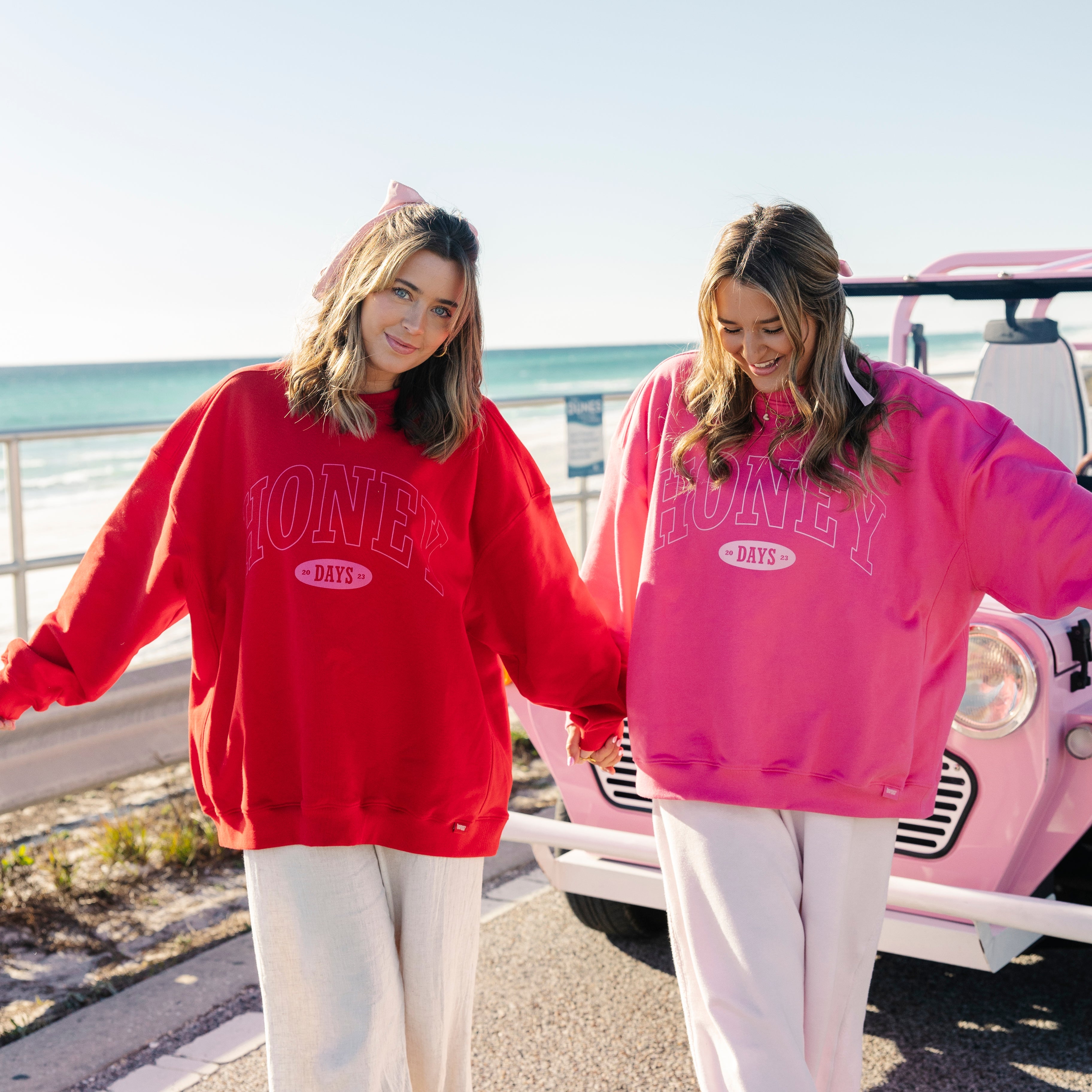 Two women smiling and holding hands by the ocean, wearing matching oversized sweatshirts—one red, one pink—with “HONEY DAYS” text, white pants, and standing beside a pink car on a sunny day.