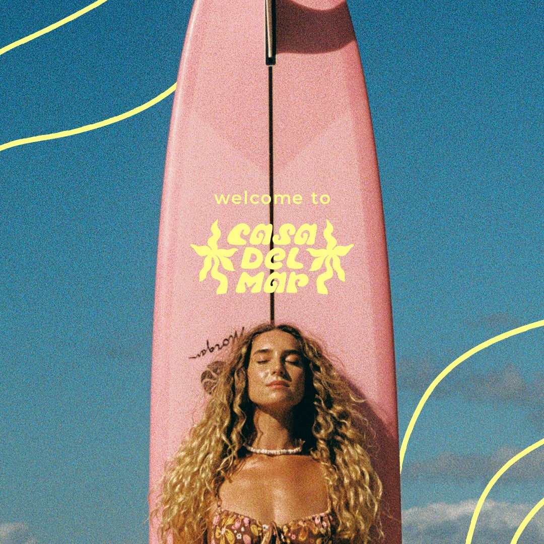 A woman in a patterned bikini stands in front of a pink surfboard with Welcome to Casa del Mar written on it. The sky is blue with light clouds and yellow wavy lines are drawn over the image.