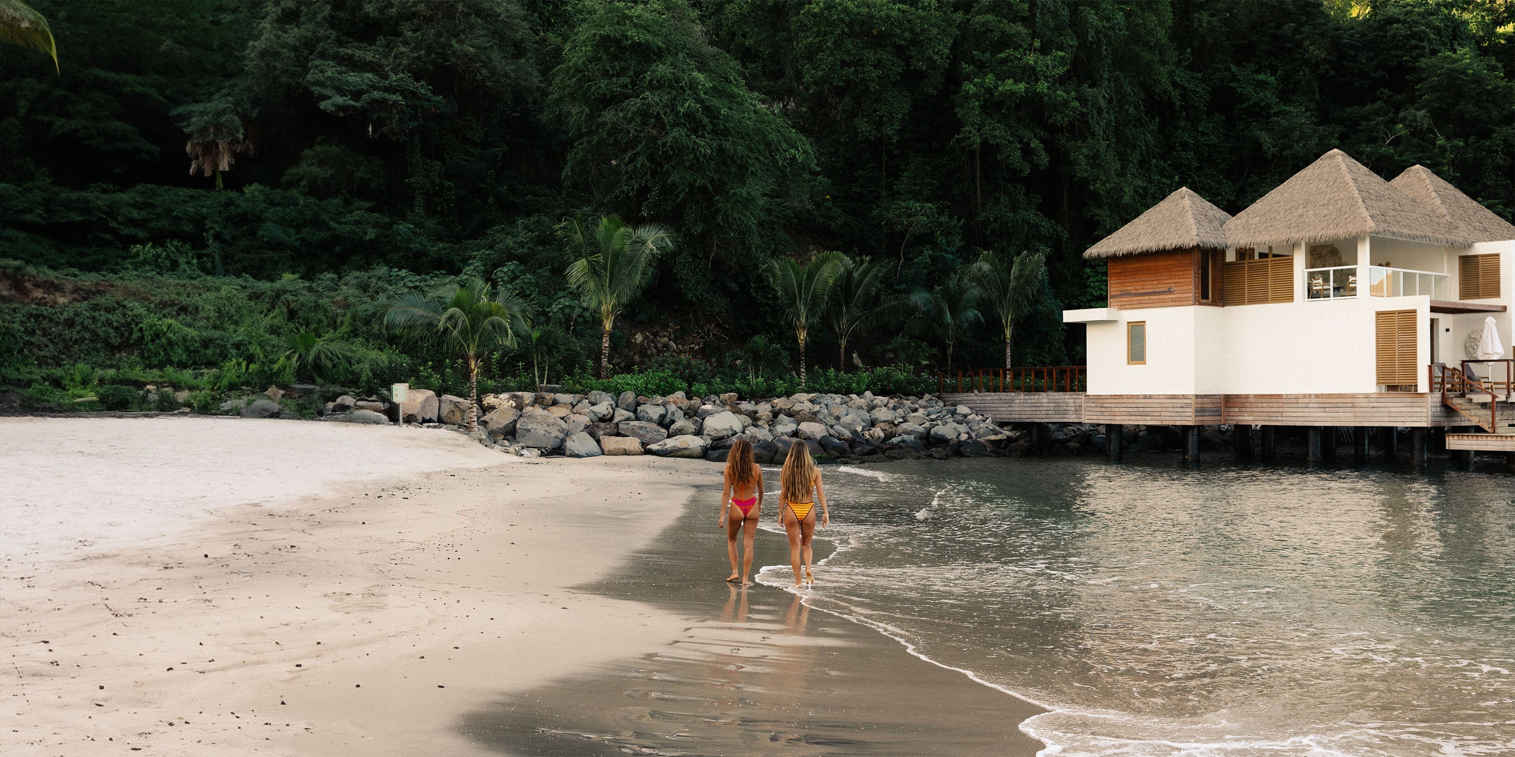 Two women in swimsuits walk hand in hand along a sandy beach toward a tropical overwater bungalow, with lush green trees in the background and calm waves at their feet.