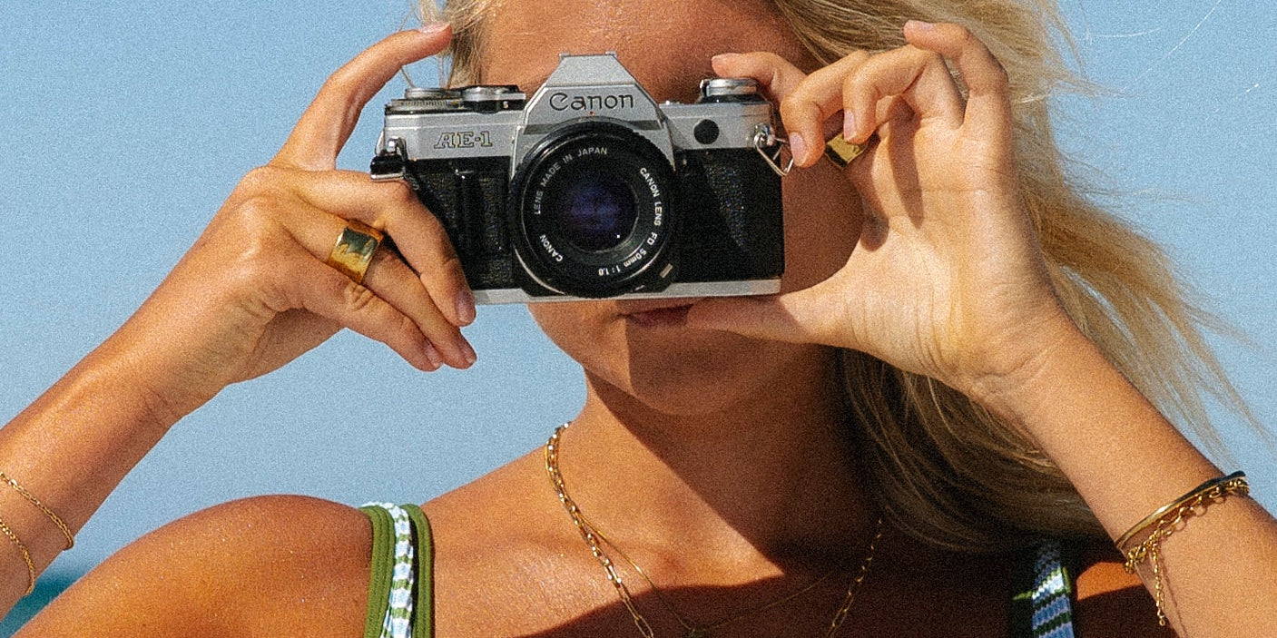 A woman wearing a yellow bikini stands outdoors among lush green tropical plants and palm trees, looking confidently at the camera.
