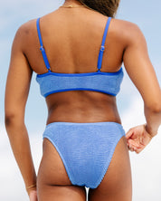 A woman stands outdoors, back to the camera, in the Coastal Blue Sporty Luxe Set by Honey Swim. The blue textured bikini contrasts with the sky as she rests her right hand near her hip.