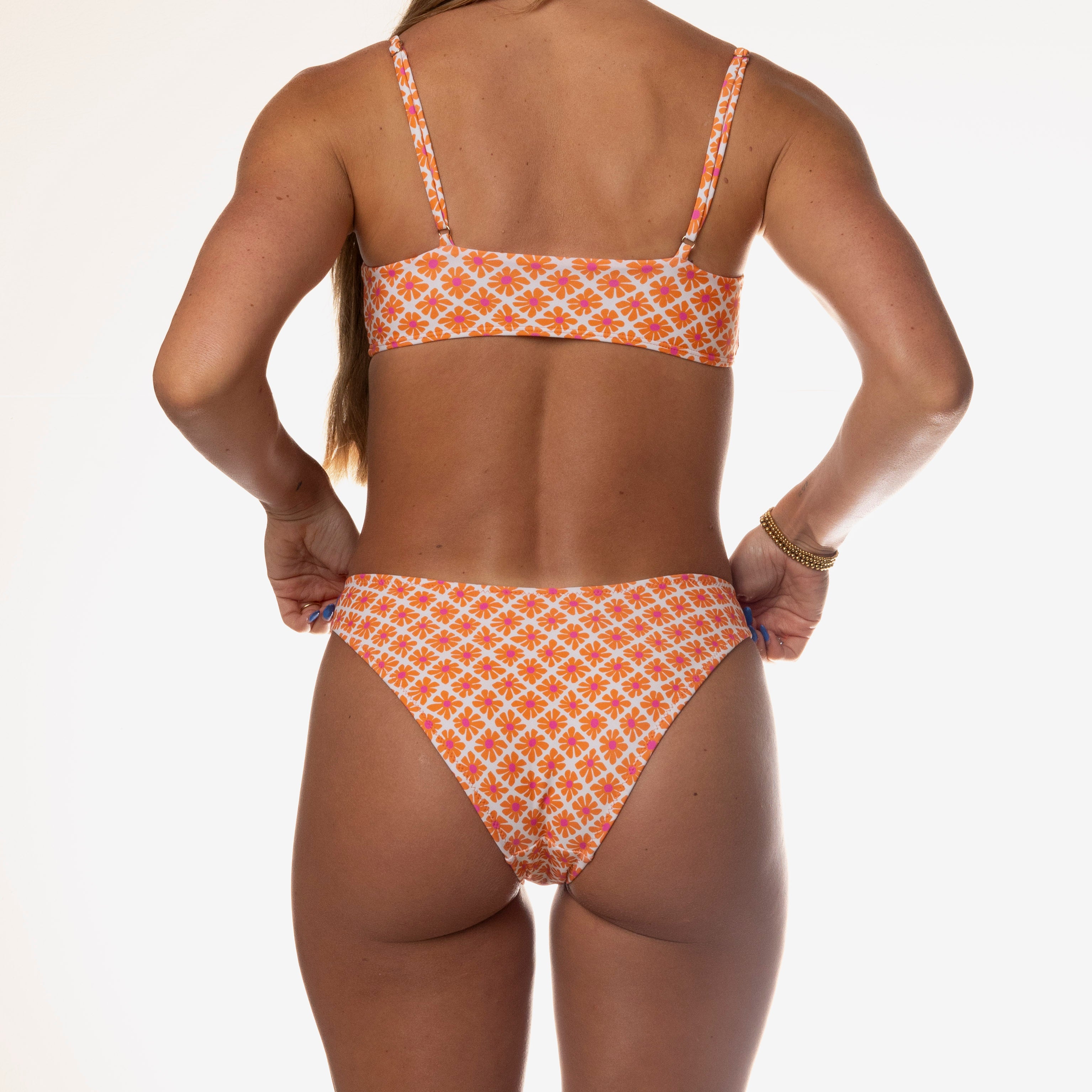 A person poses in the Honey Swim Kora Bralette Set, showcasing its timeless silhouette with an orange and white floral pattern bikini against a plain white background, highlighting the back view.