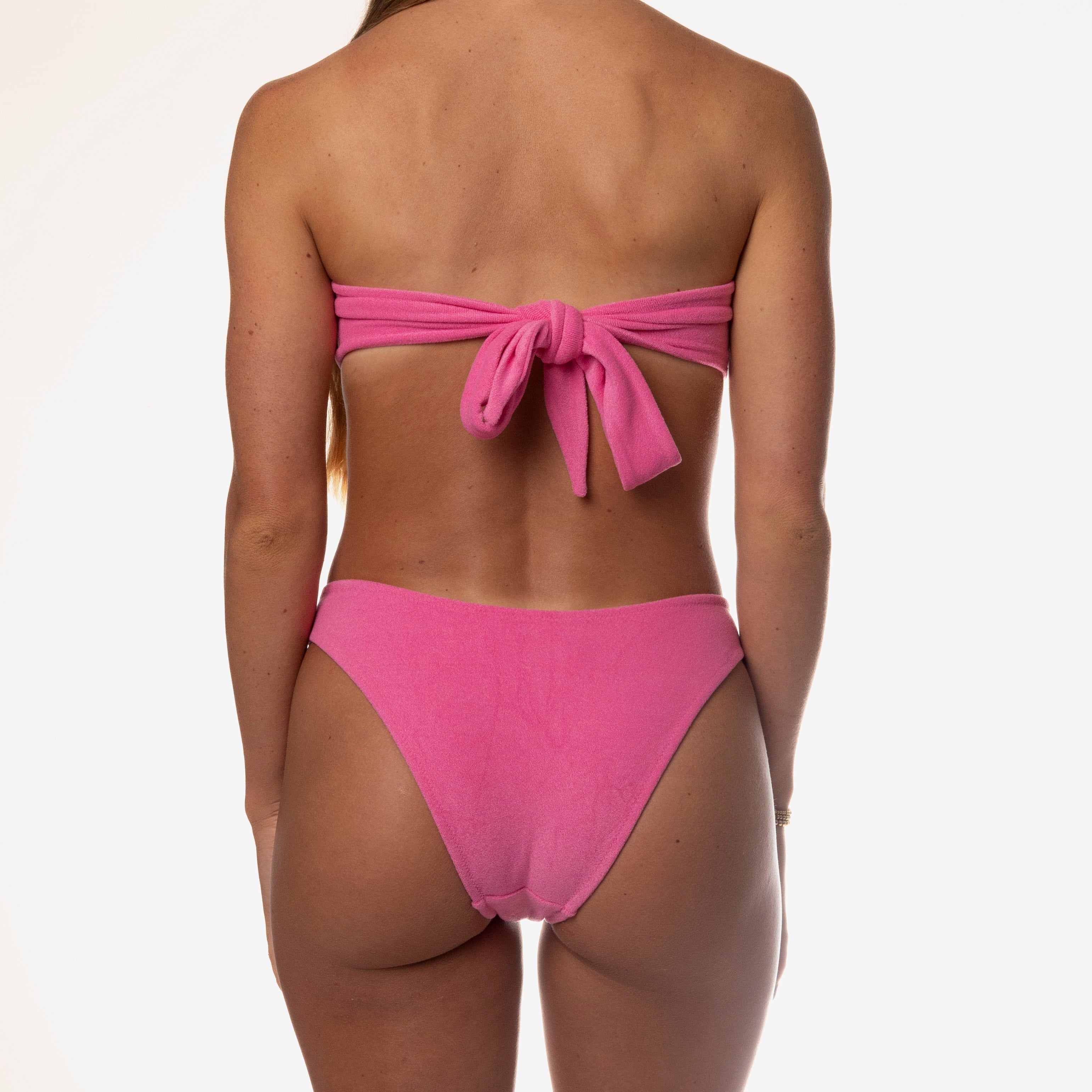 Against a white backdrop, someone wears the Flirty Terry Bandeau Set by Honey Swim, a playful pink bikini featuring a back bow, ideal for sunny days.