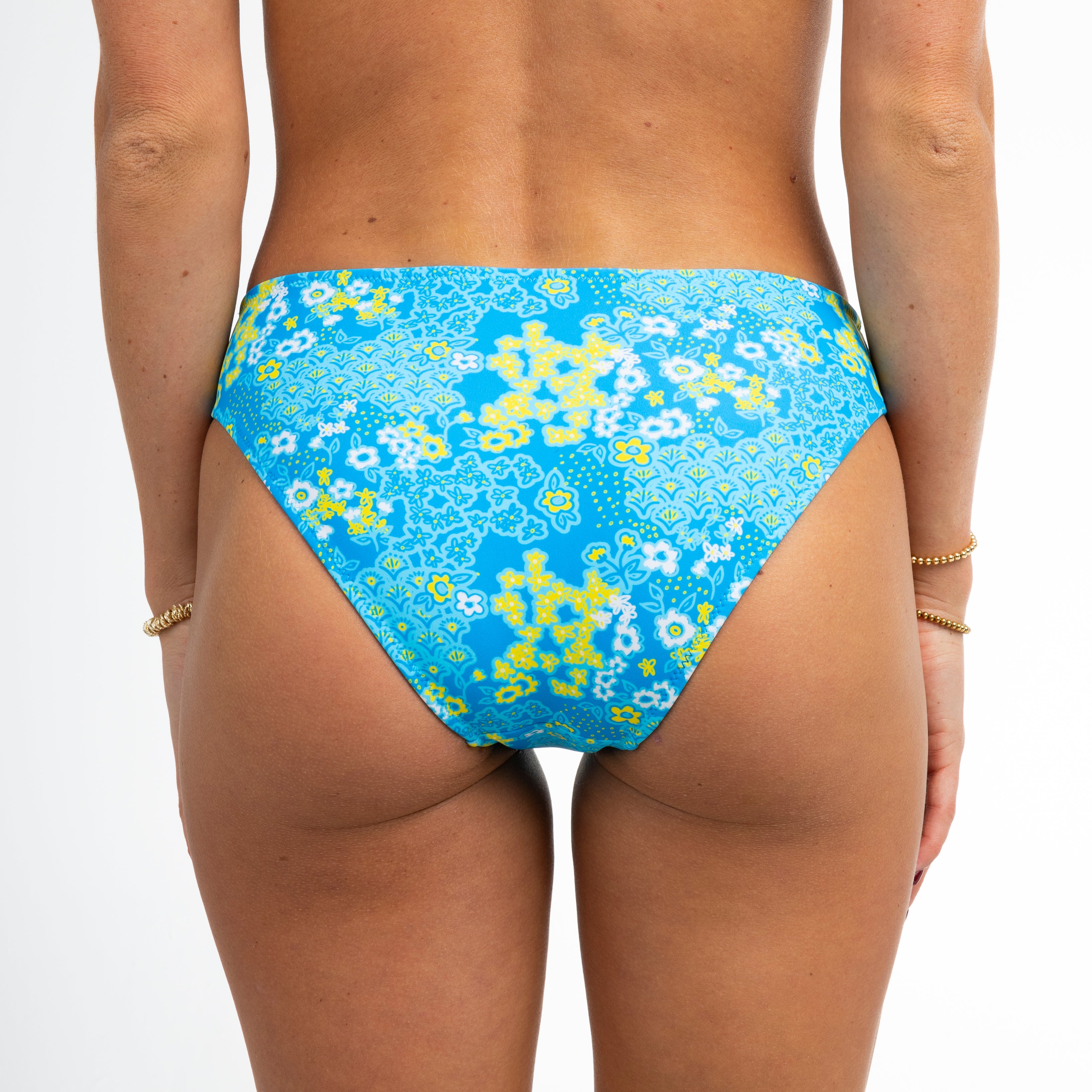 A person showcases the back of Honey Swims Blossom bikini bottoms, featuring an eclectic pattern of yellow and white florals. Their relaxed hands wear two gold bracelets each against a plain light backdrop.