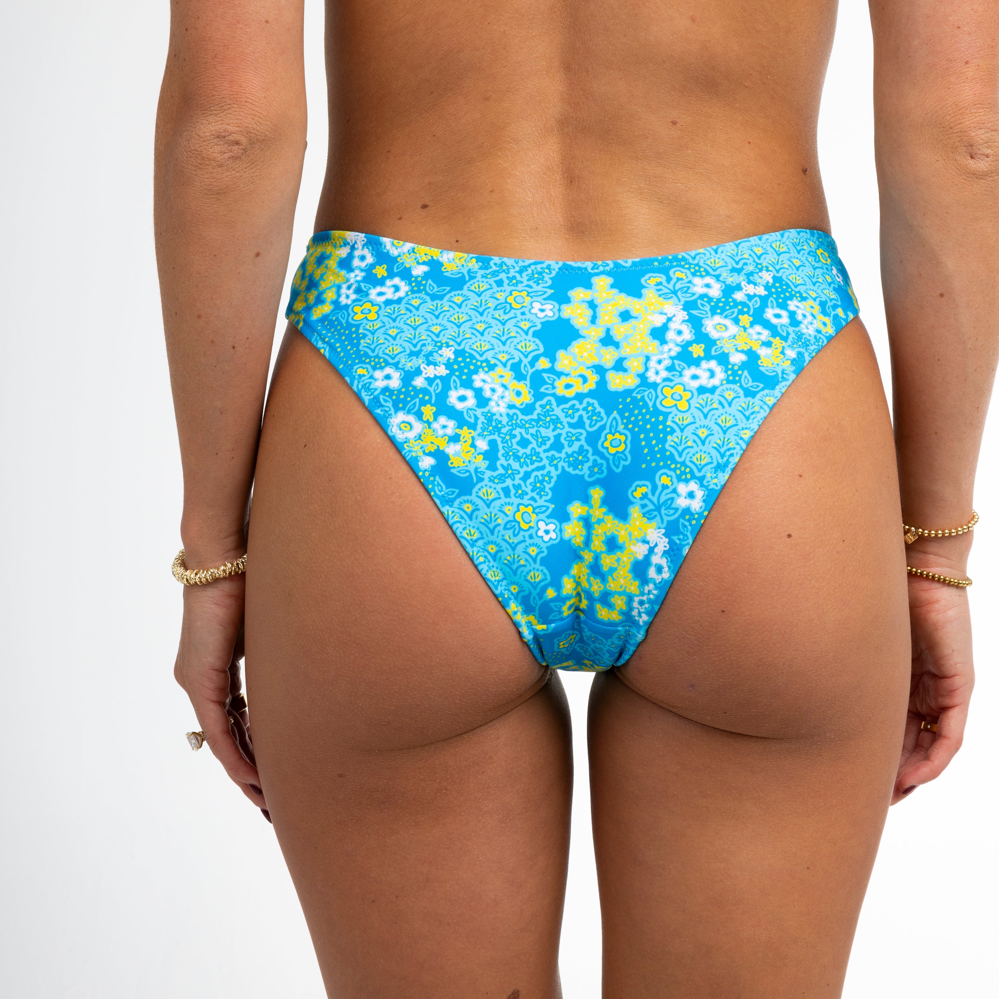 A person models the Blossom bikini bottoms by Honey Swim, featuring an eclectic floral pattern against a white background. Gold bracelets on their hands highlight the complimentary colors beautifully.