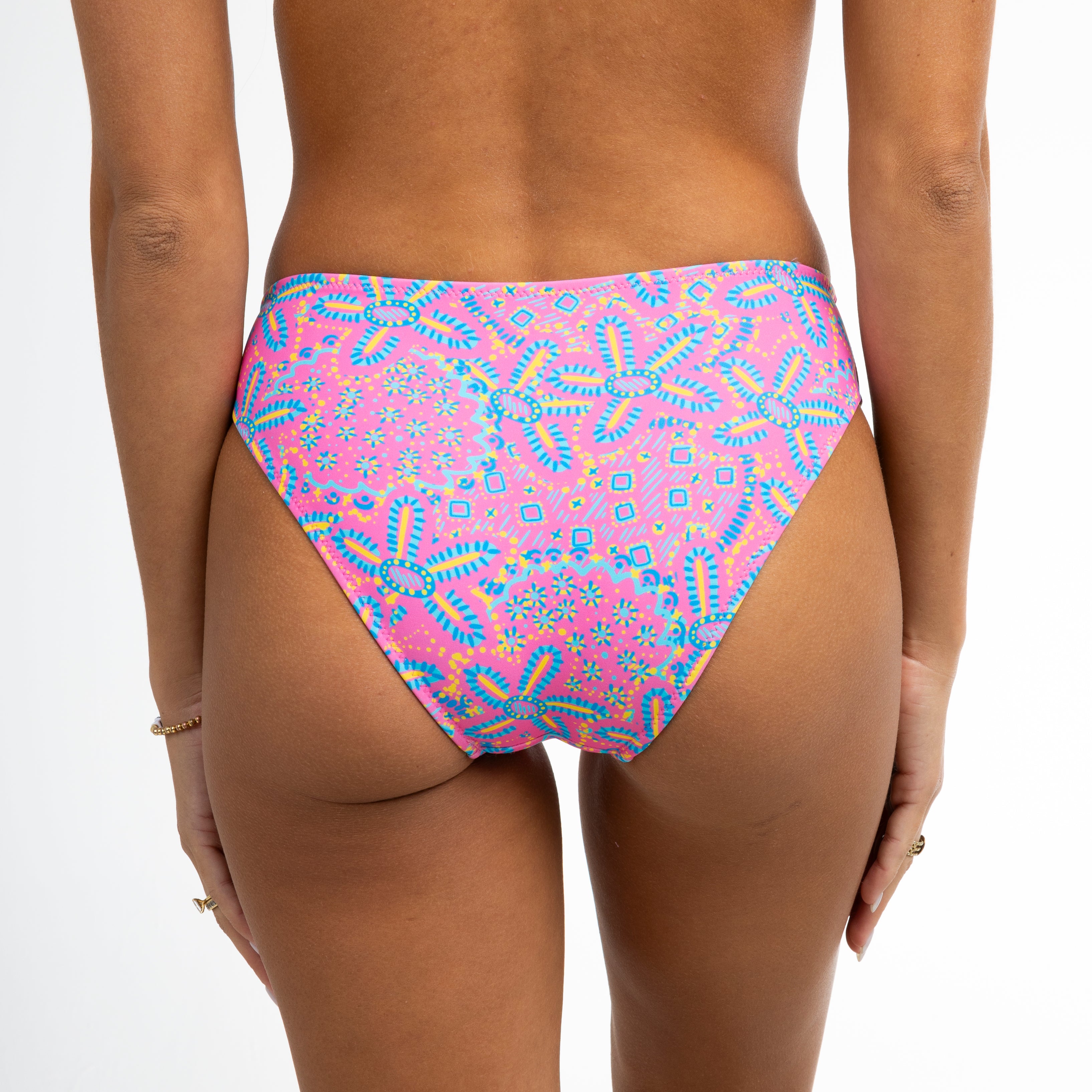 A person wears Honey Swims Starfish full coverage bottoms in a whimsical multicolored abstract pattern, paired with thin bracelets on their left wrist, against a white background with their back to the camera.