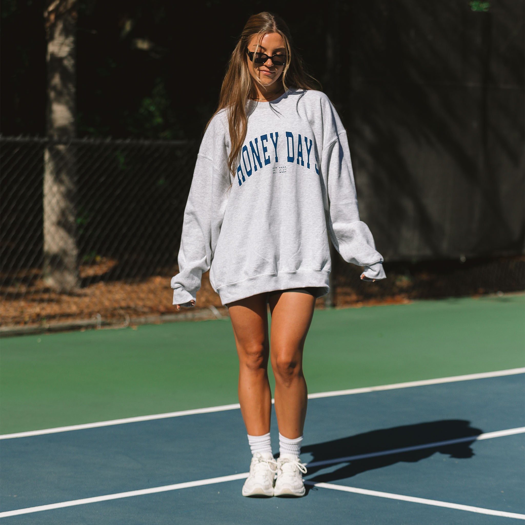 A person stands on a tennis court, wearing sunglasses, white socks, sneakers, and the Honey Days Crewneck by Honey Swim. With long hair and a relaxed stance, they embody an everyday comfy look while gazing at the camera.