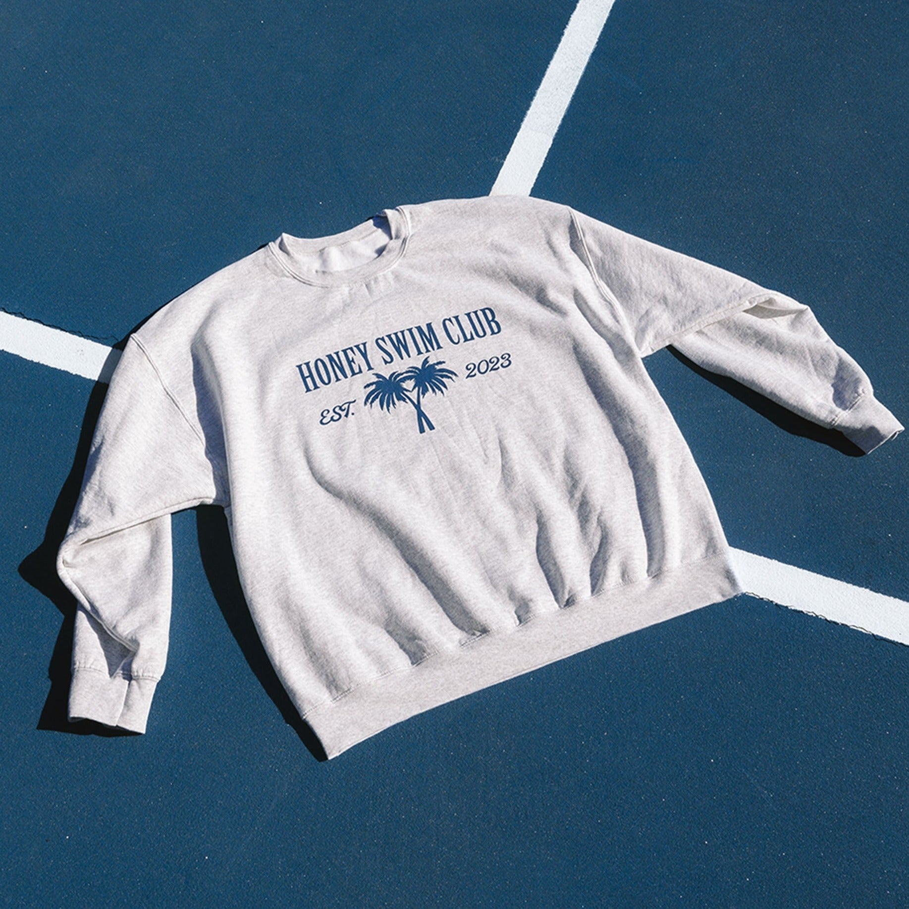 The Honey Swim Palm Crewneck, a gray oversized sweatshirt with Honey Swim Club and Est. 2023 in blue and a palm tree graphic, is styled on a dark blue background with intersecting white lines.