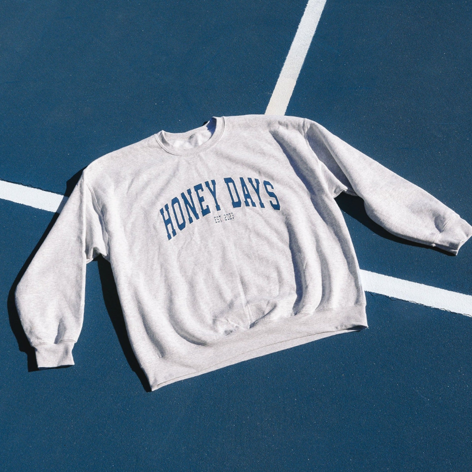 The Honey Swims Honey Days Crewneck is a gray oversized sweatshirt featuring HONEY DAYS and EST. 2020 in dark blue on the chest, laid flat on a blue sports court, offering a comfy fit for casual outings.