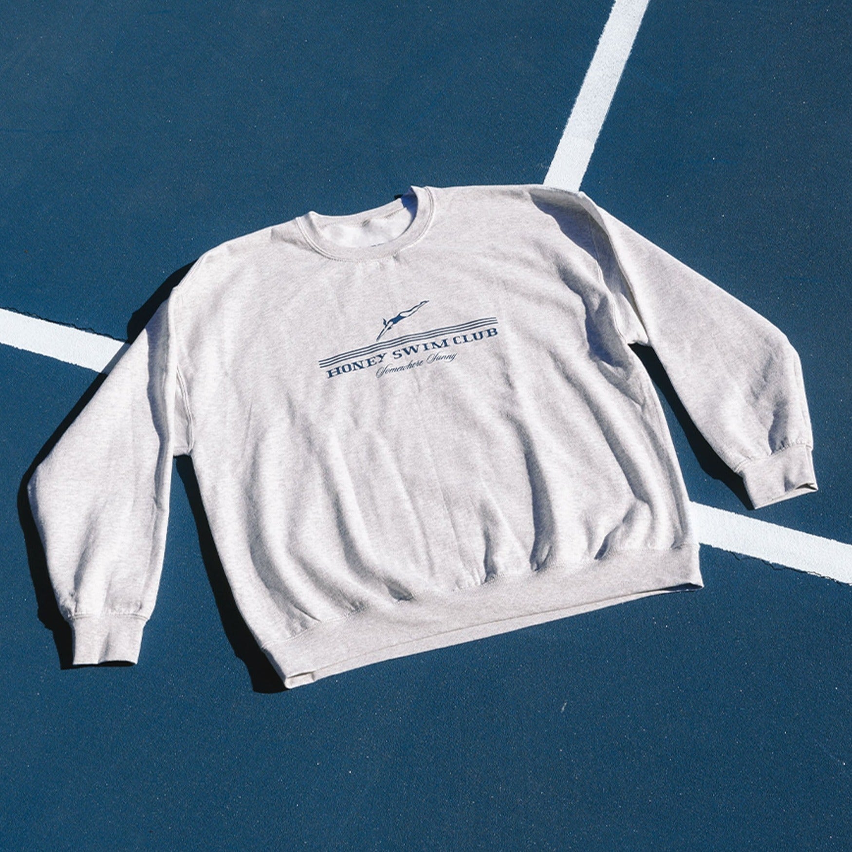 The Somewhere Sunny Crewneck by Honey Swim is an oversized gray sweatshirt with a front graphic design. Its shown on a blue background with white lines resembling a sports court, perfect for that comfy fit vibe.