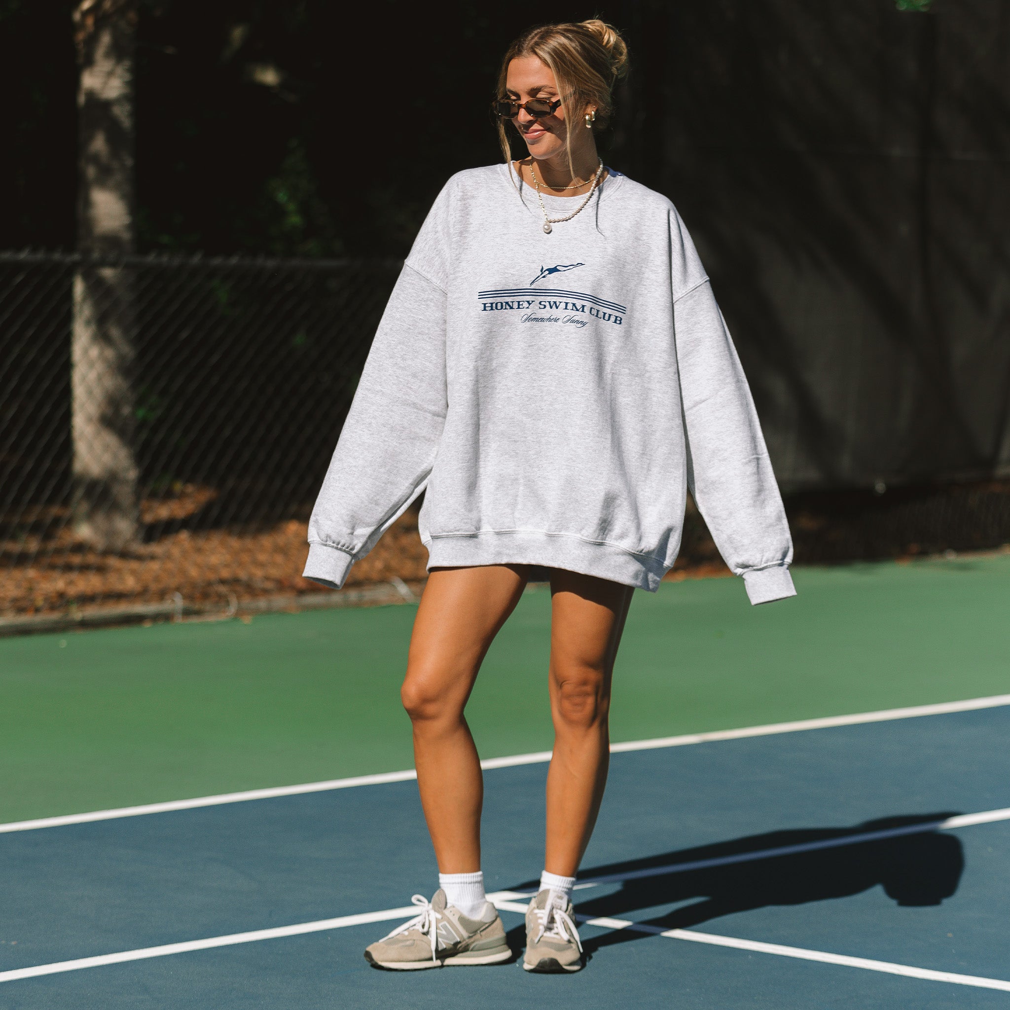 A woman wearing sunglasses stands on a tennis court in an oversized Somewhere Sunny Crewneck by Honey Swim, paired with shorts and sneakers. Her hair is styled in a playful bun as she smiles, framed by dark fences and trees.