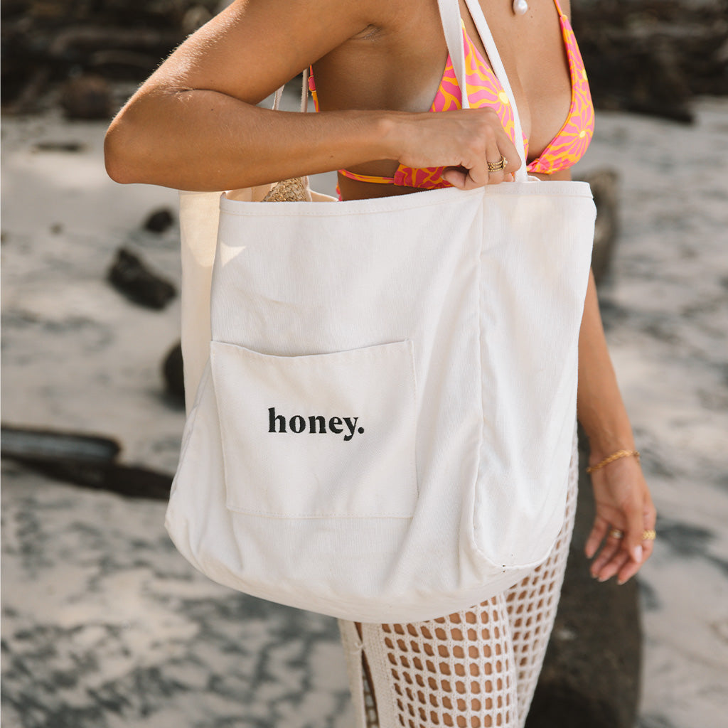 On the beach, someone carries a Honey Swims Honey Tote Bag while wearing a colorful bikini top and white mesh cover-up skirt; the totes canvas contrasts beautifully with the sandy and rocky backdrop.