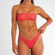 A woman models the Honey Swim Azalea Cinched Bandeau Set, featuring a white floral print with pink blooms and no-tan lines, standing against a plain white background with one hand on her hip.