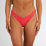 A woman models the Honey Swim Azalea | Classic Bottoms in red with a white floral print, standing against a plain white background and accessorized with bracelets and rings—ideal for beach days.