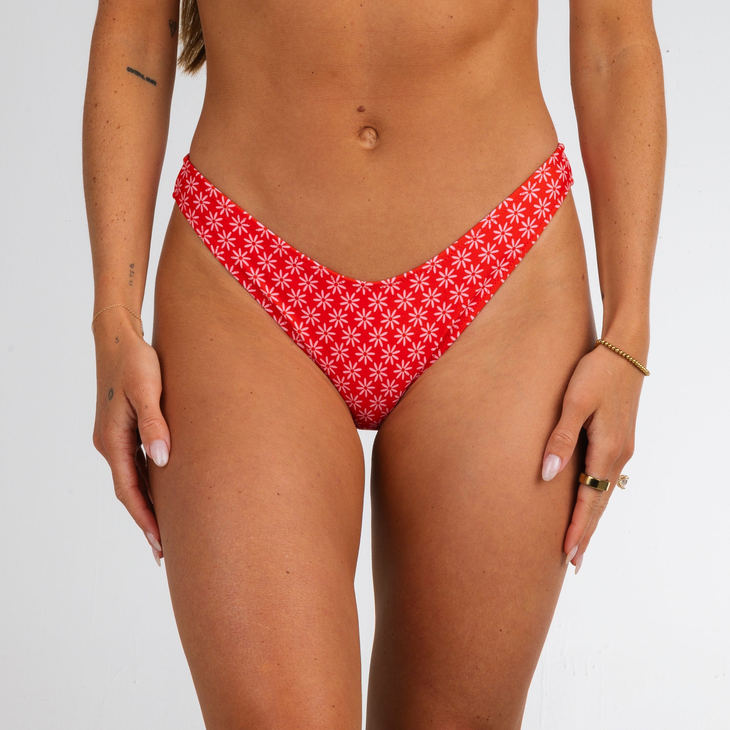 A woman models the Honey Swim Azalea | Classic Bottoms in red with a white floral print, standing against a plain white background and accessorized with bracelets and rings—ideal for beach days.