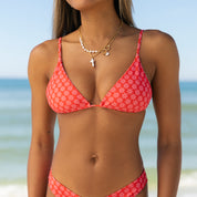 A woman in the Honey Swim Azalea Triangle Set with adjustable straps and layered necklaces stands on a beach, ocean and blue sky in the background.