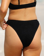 A close-up of a person wearing honeyswim’s Black Luxe | Classic Bottom bikini with moderate coverage on a beach, showing only the midsection and upper legs, hand resting at the side.