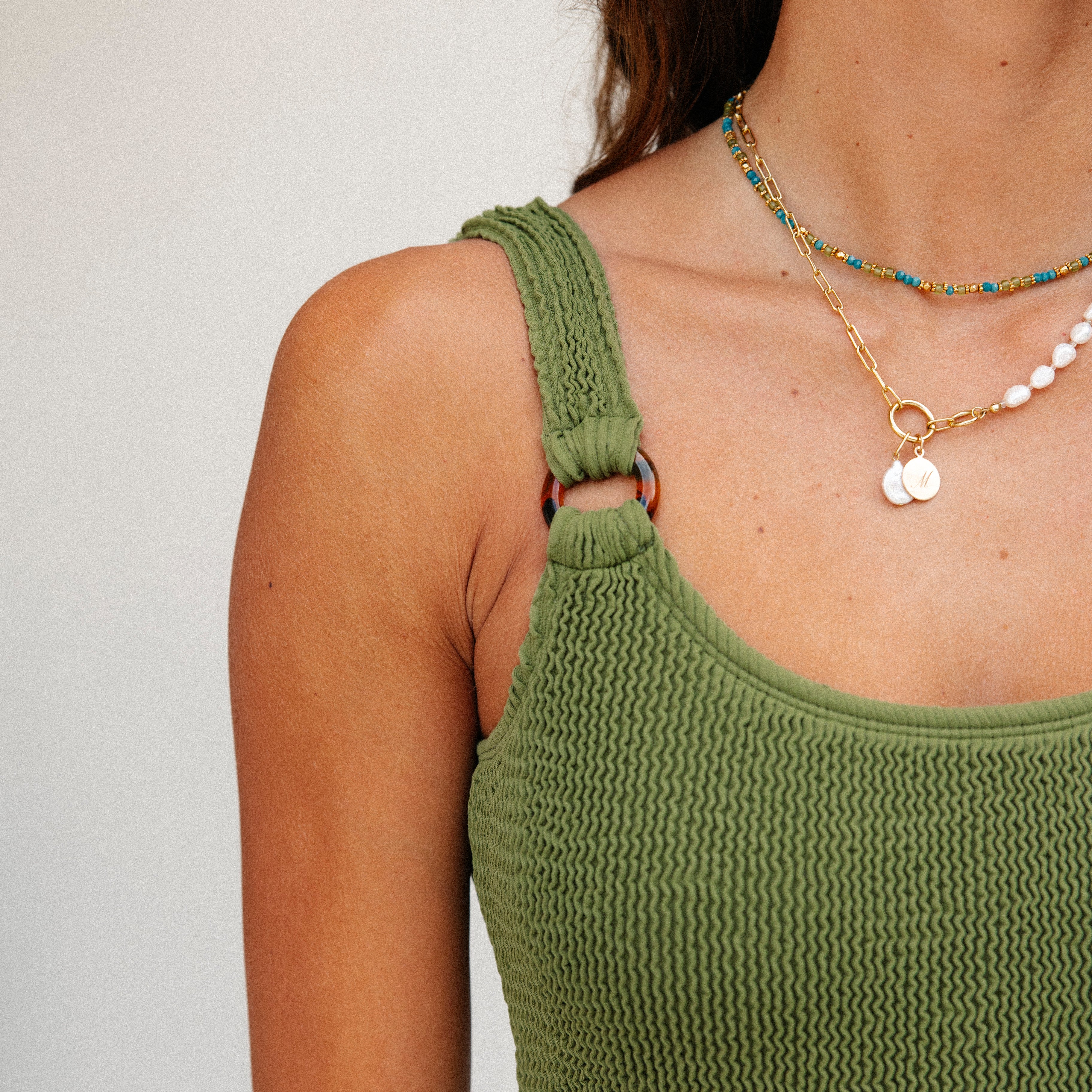 A person models the Olive Luxe One Piece by Honey Swim, featuring ring details and a perfect cut. Layered necklaces with colorful beads and small pendants add elegance against a neutral background, enhancing the soft allure of this ensemble.
